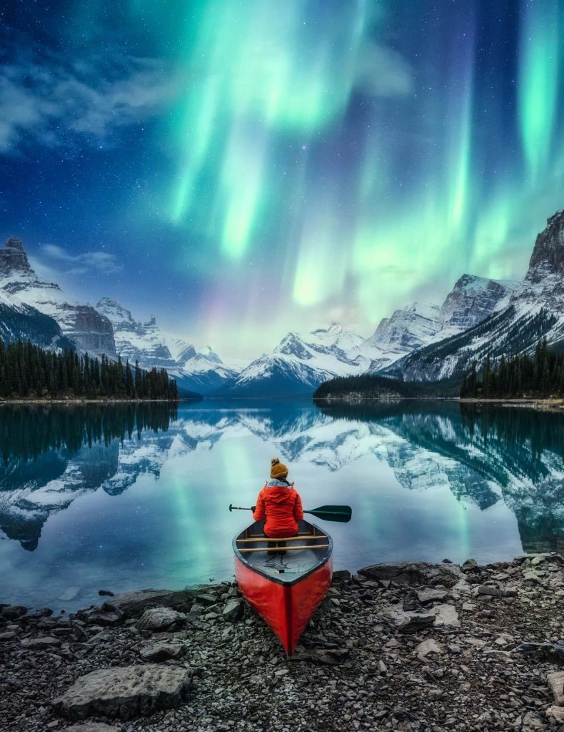a person sitting in a canoe in a lake surrounded by mountains and northern lights in the sky